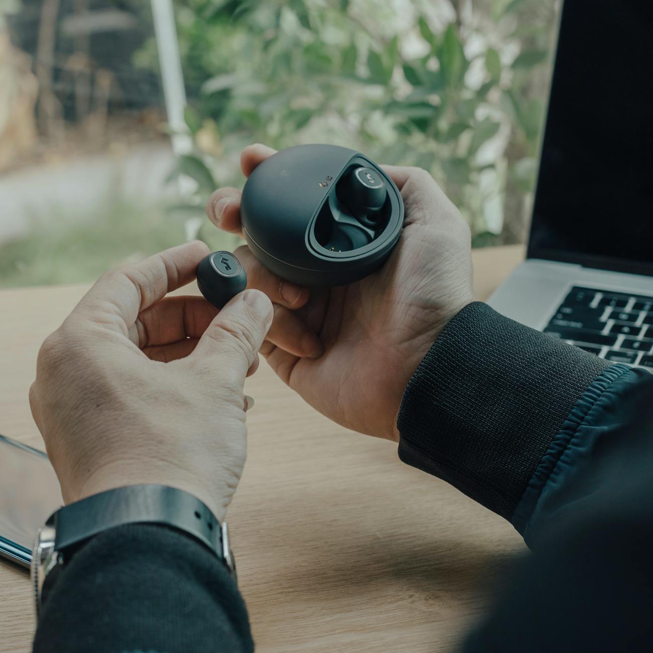 Unrecognizable male sitting at wooden table with laptop and holding true wireless earphones placed in charging case